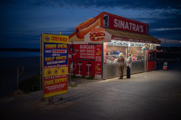 A couple of people standing outside a hot dog stand.