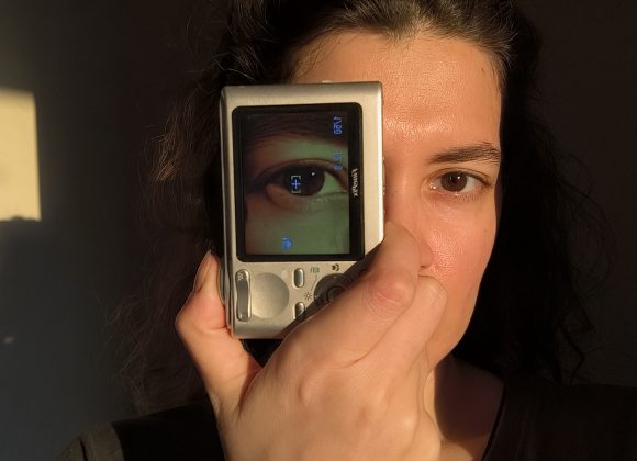 A person holds a camera displaying their eye over one side of their face in warm sunlight.
