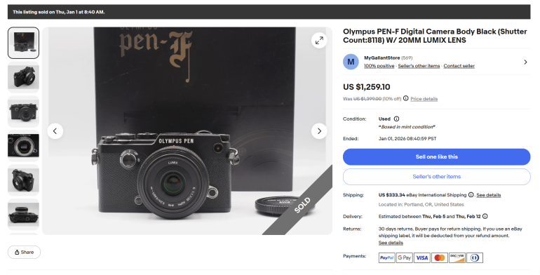 Black Olympus PEN-F camera with 20mm Lumix lens shown in front of its box; listing marked as sold.