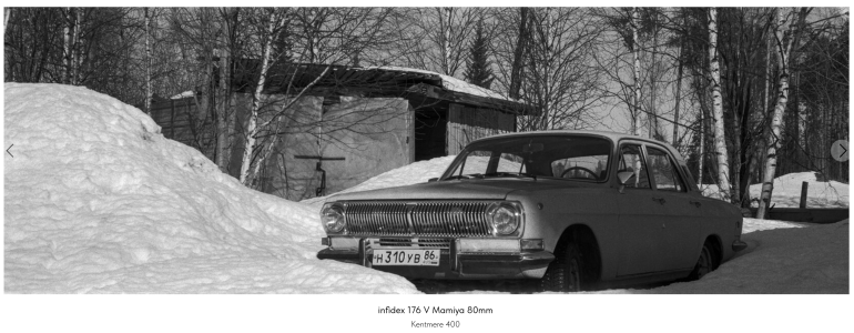 A vintage car is parked in deep snow beside a wooden shed and leafless trees in winter.