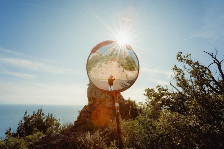 A round mirror reflects a person standing on a path, with sunlight shining above and trees surrounding the area.