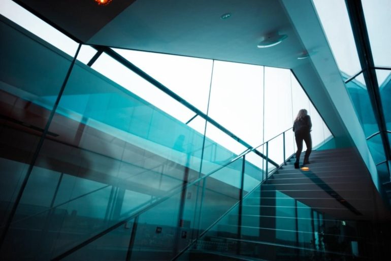 A person stands on a modern glass staircase inside a building with large, slanted windows.
