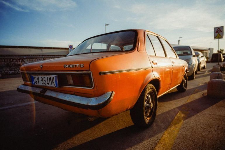 An old orange Opel Kadett car parked on a street in sunlight, with another car beside it.