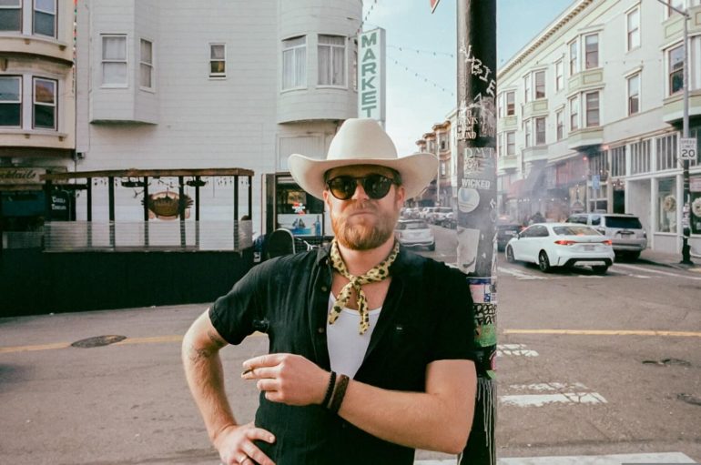 Man in a cowboy hat and sunglasses stands on a city street, holding a cigarette and posing confidently.