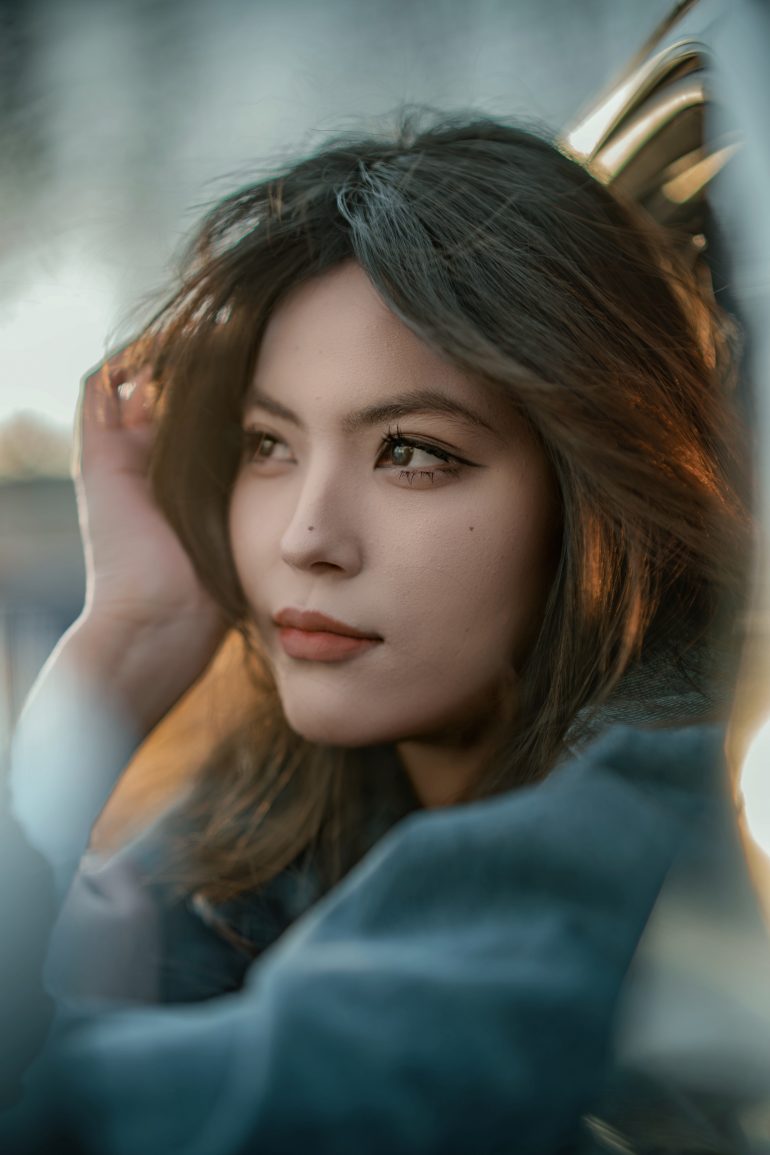 A woman with long dark hair gazes thoughtfully into the distance, hand touching her hair.