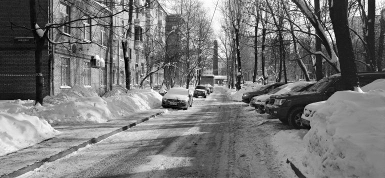 Snow-covered cars line a narrow city street with buildings and bare trees on a sunny winter day.