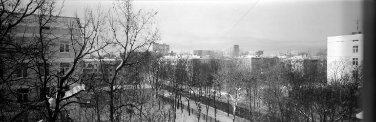 Black and white cityscape with leafless trees, snowy ground, and several apartment buildings in the background.