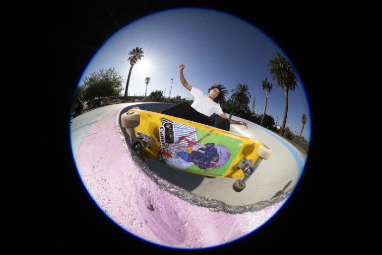 A skateboarder does a trick on a ramp, captured with a fisheye lens under a sunny sky with palm trees.