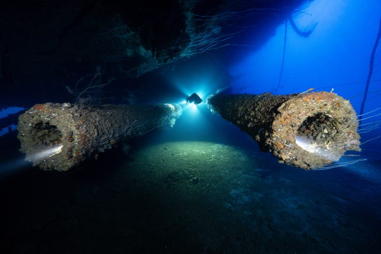 A scuba diver explores beneath a sunken shipwreck, illuminated by blue light and surrounded by debris.