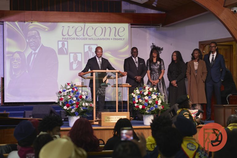 A pastor and his family stand on stage as a congregation welcomes them with flowers and a slideshow background.