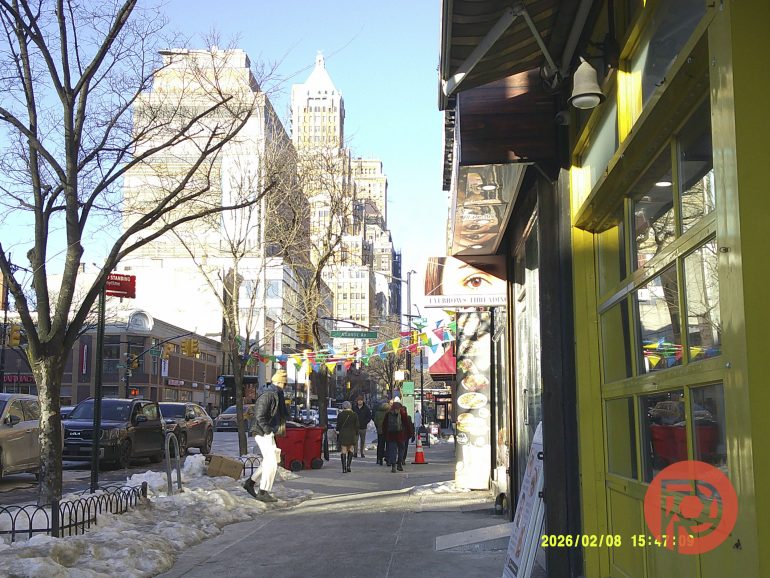 Busy city sidewalk with people walking, colorful flags, and tall buildings under a clear blue sky.