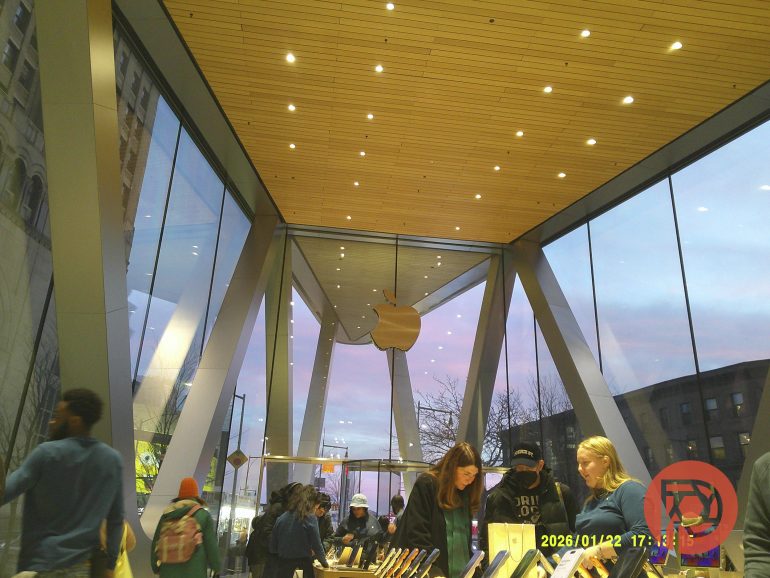 People browsing devices inside an Apple Store with large glass windows and a sunset sky outside.