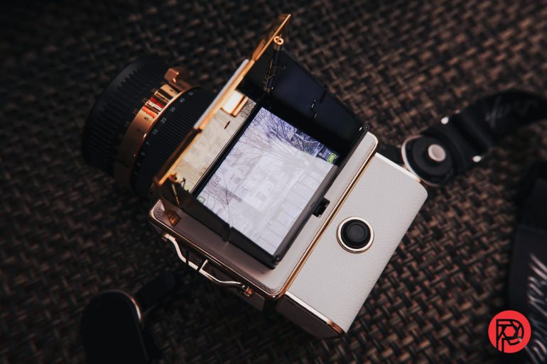 A vintage camera with a flip-up viewfinder displays a tree-lined street; camera strap and logo visible nearby.
