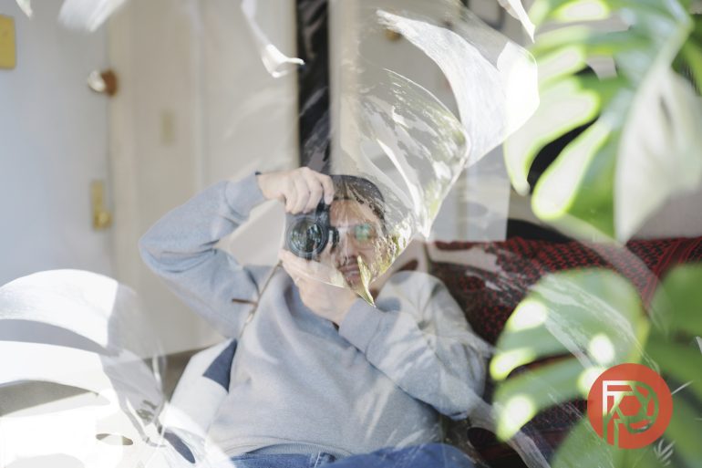 Person in gray sweater takes a photo indoors, partially obscured by large green leaves and reflections.