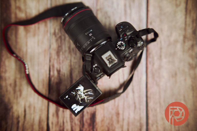A Canon camera with a strap rests on wood, its screen flipped out showing a close-up image of a flower.