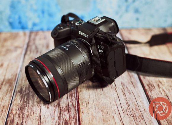 A Canon EOS R6 camera with a lens rests on a wooden surface with a blue background.