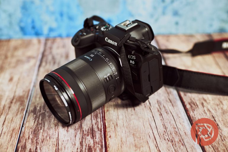 A Canon EOS R6 camera with a lens rests on a wooden surface with a blue background.