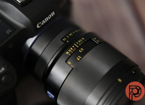 Close-up of a Canon camera with a lens showing focus and aperture markings, on a blurred background.