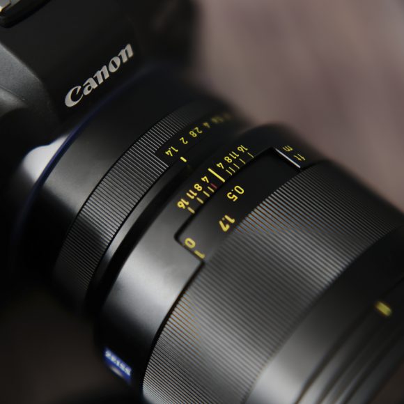 Close-up of a Canon camera with a lens showing focus and aperture markings, on a blurred background.