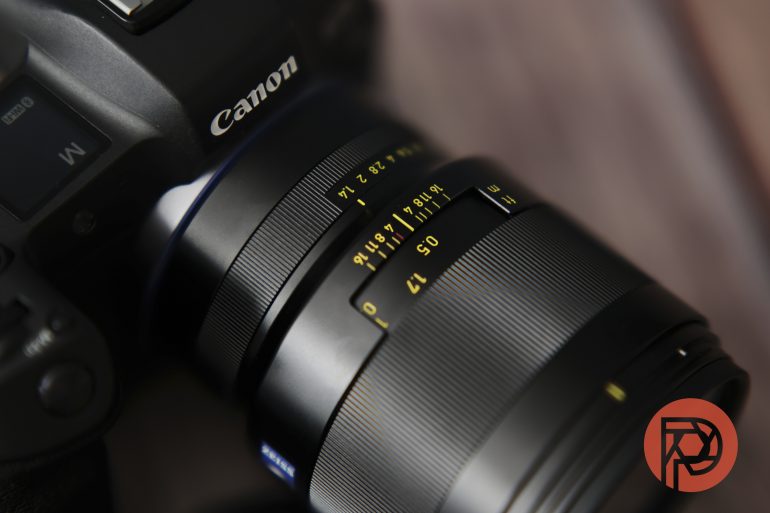Close-up of a Canon camera with a lens showing focus and aperture markings, on a blurred background.