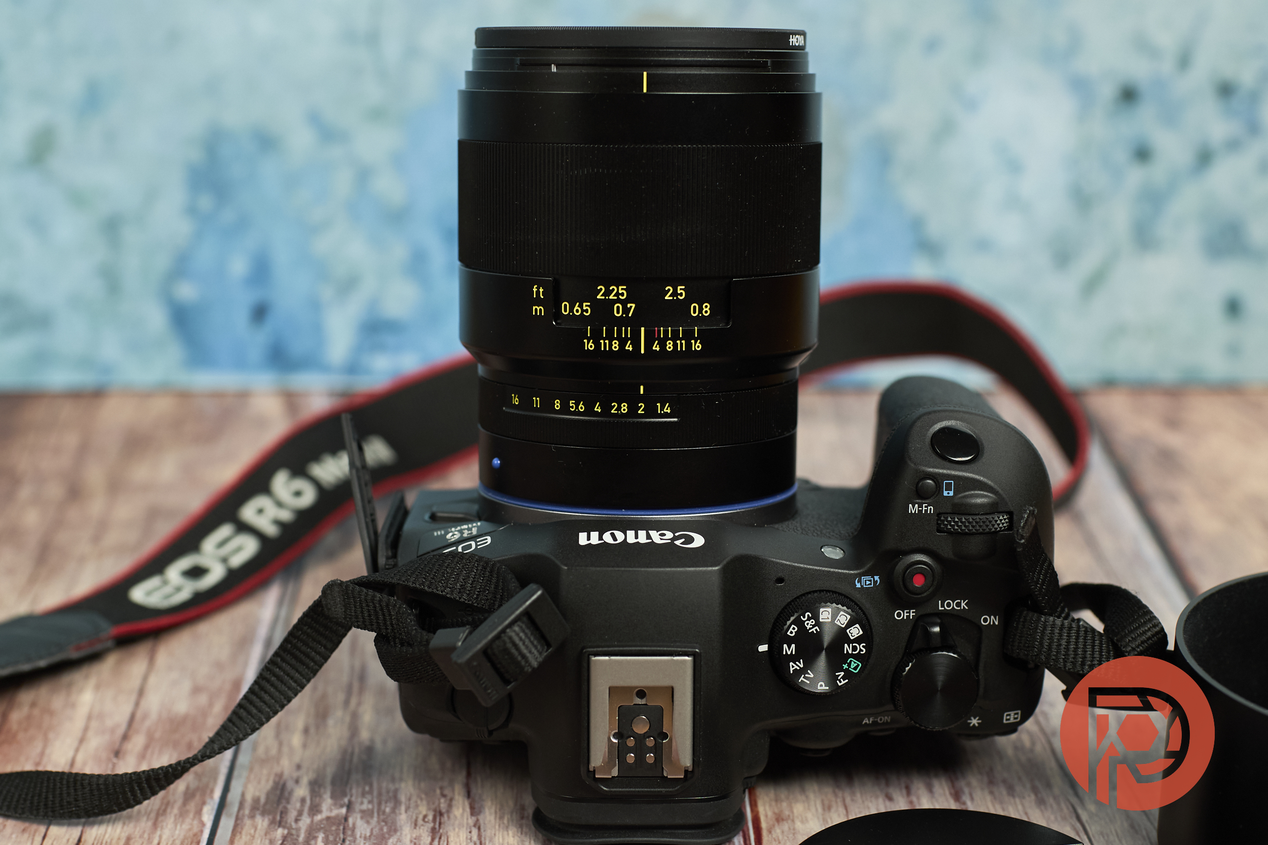 Canon EOS R6 camera with a black lens on a wooden surface, camera strap visible, and blue background.