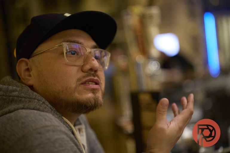 A person in glasses and a cap gestures while talking in a dimly lit indoor setting.
