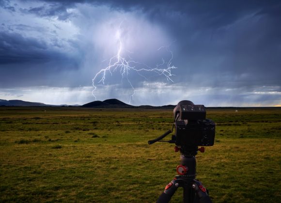 A camera on a tripod captures lightning striking over grassy plains and distant hills under a stormy sky.