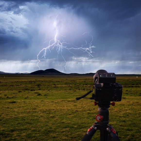 A camera on a tripod captures lightning striking over grassy plains and distant hills under a stormy sky.