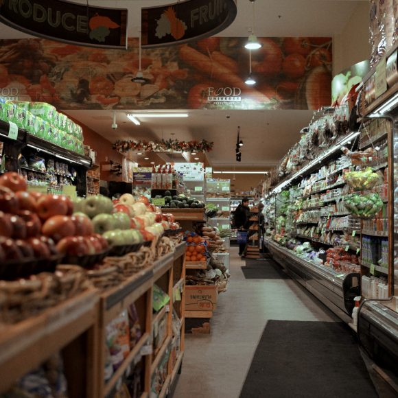 A grocery store aisle with produce on the left and refrigerated items on the right. Shoppers are in the background.