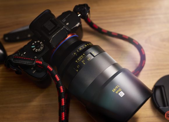 A close-up of a camera with a large lens and a red-and-black braided strap on a wooden surface.