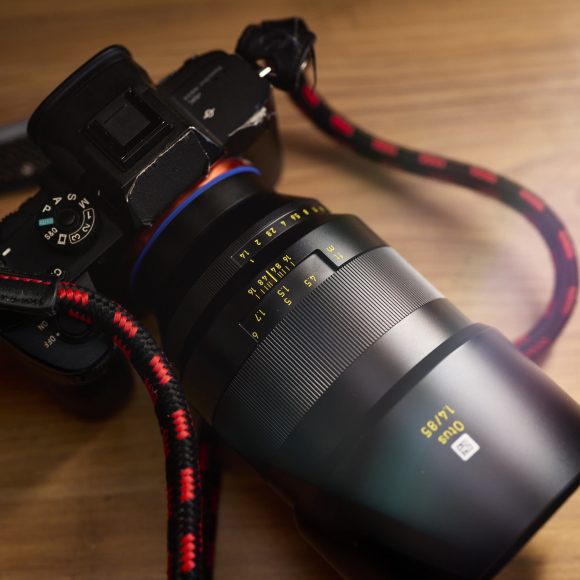 A close-up of a camera with a large lens and a red-and-black braided strap on a wooden surface.