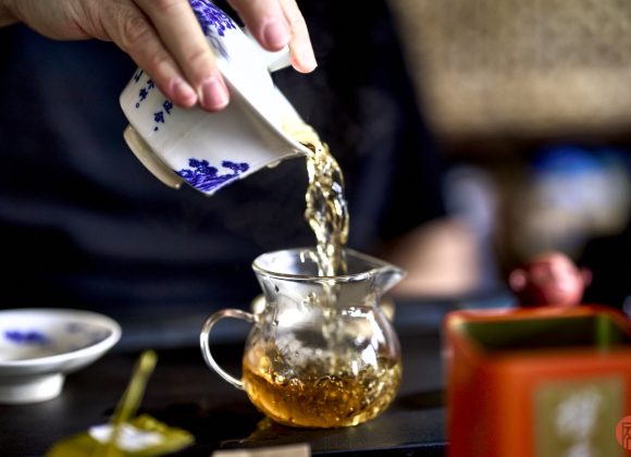 Hand pouring tea from a blue and white cup into a glass pitcher during a traditional tea ceremony.