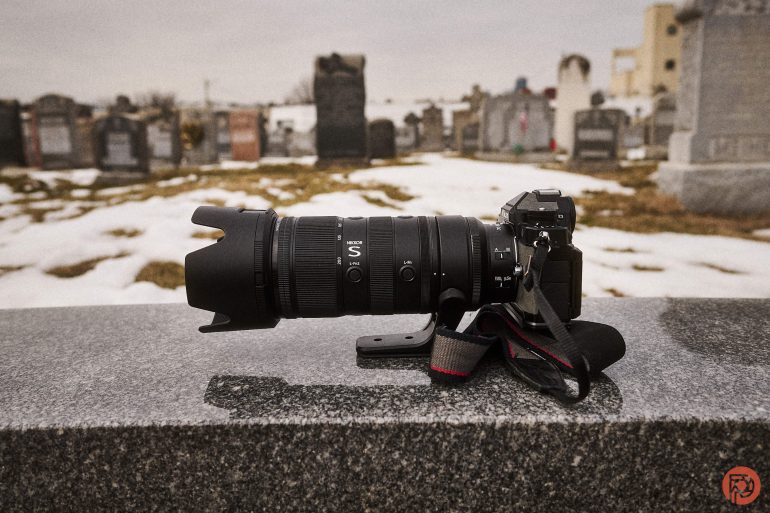 A camera with a large lens rests on a stone surface in a snowy cemetery with gravestones in the background.
