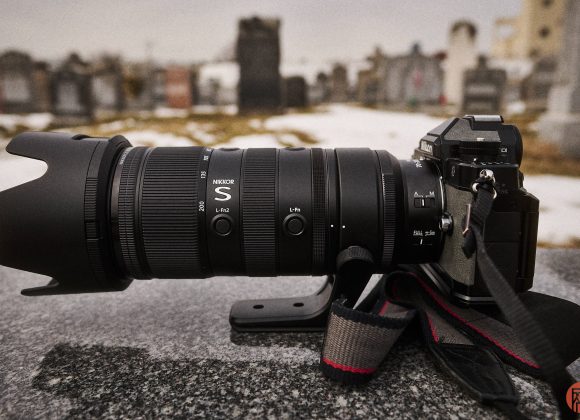 A black DSLR camera with a large telephoto lens sits on a stone surface in a cemetery.