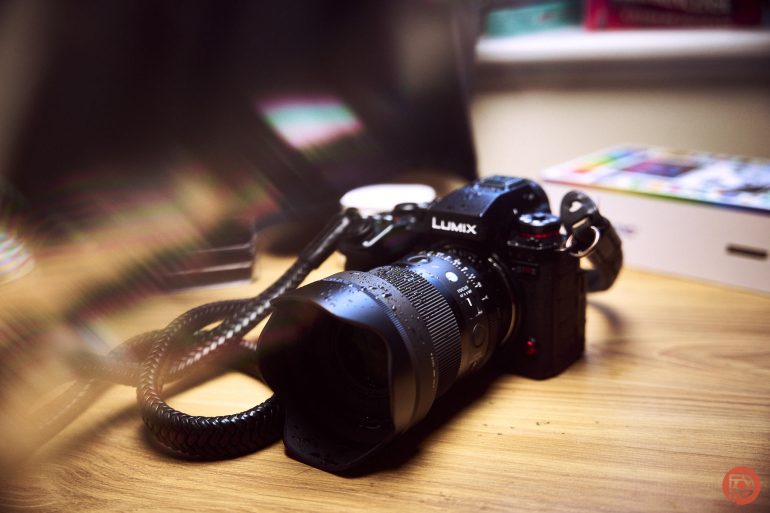 A Lumix camera with a lens and strap rests on a wooden desk beside a colorful box.