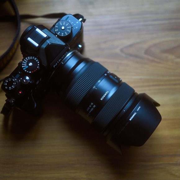 A black digital camera with a large zoom lens sits on a wooden surface in soft lighting.