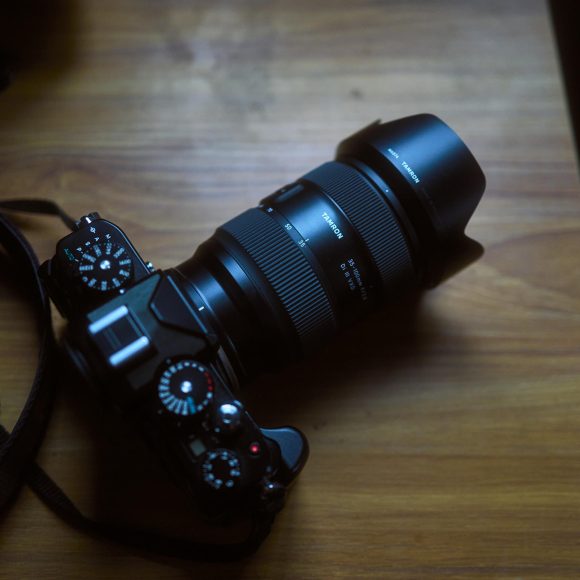 A black DSLR camera with a zoom lens sits on a wooden surface in dim lighting.