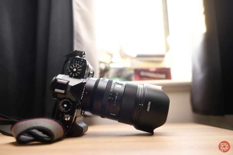 A black Fujifilm camera with a Viltrox lens rests on a wooden surface near a window.