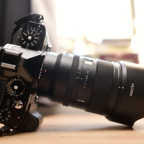 A black digital camera with a Viltrox lens rests on a wooden surface in soft natural light.