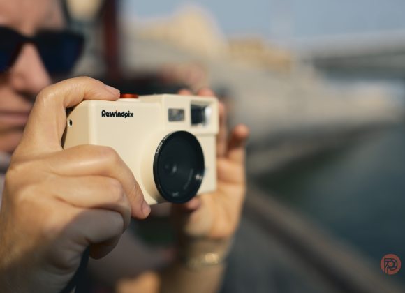 A person holding a white Rewindpix camera outdoors, with blurred background and sunlight.