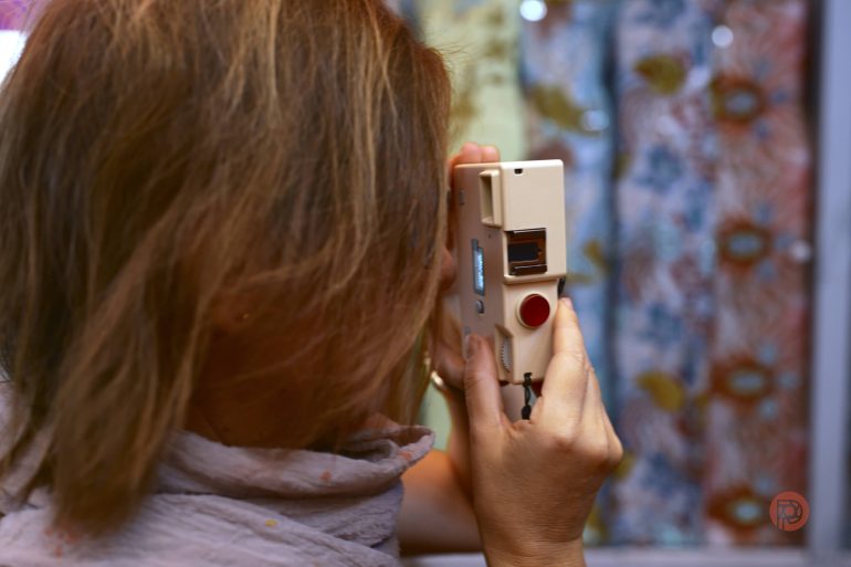 A person holds a vintage camera up to their eye, facing a colorful patterned background.