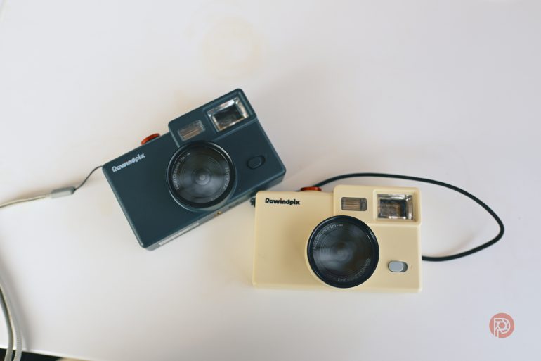 Two retro-style film cameras, one blue and one cream, placed on a white surface with wrist straps attached.