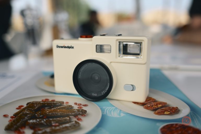 A white vintage camera sits on a table, surrounded by plates of food with red garnish.