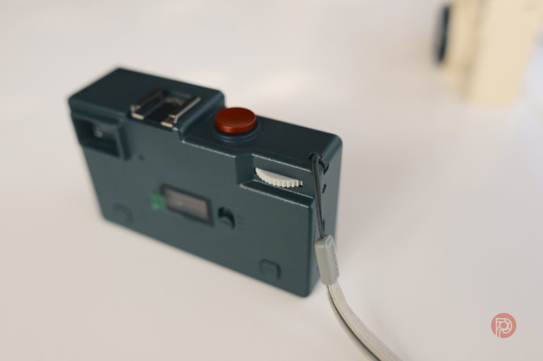 Teal compact camera with a red shutter button and a wrist strap, placed on a light surface.