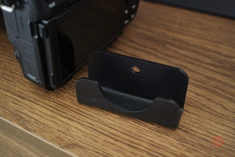 A black metal wall mount bracket sits on a wooden surface next to a digital camera.