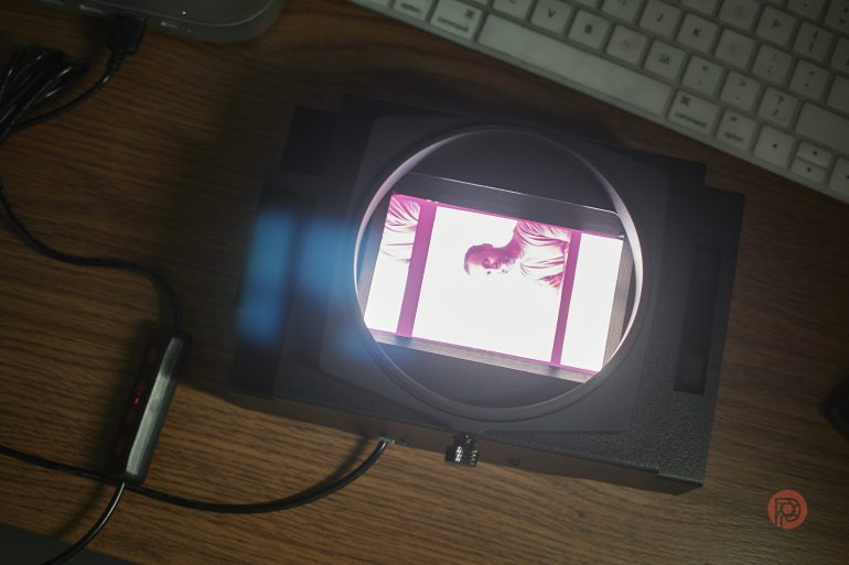 A film negative is viewed through a magnifier on a wooden desk with a keyboard nearby.