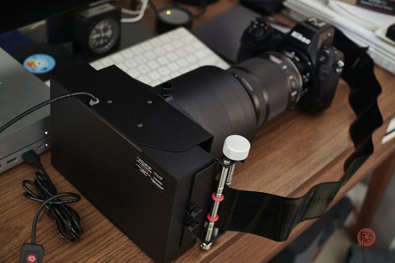 A camera with a large lens attached to a black scanning device on a wooden desk with electronics nearby.