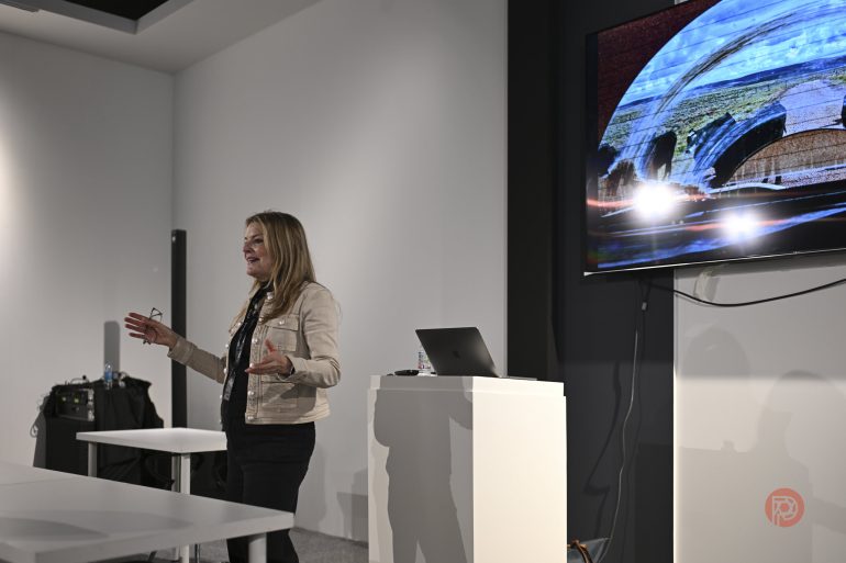 A woman gives a presentation next to a screen displaying abstract art in a modern conference room.