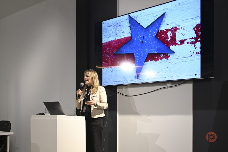 A woman speaks at a podium with a microphone; a screen shows a blue star on a red and white background.
