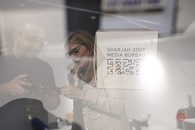 Two people examine a camera near a Sharjah Govt Media Bureau sign; image has a soft, double-exposure effect.